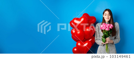 Valentines day. Romantic pretty woman close eyes and smelling beautiful flowers, standing near heart balloons, blue background 103149461