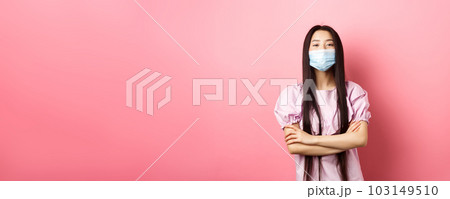 Covid-19, pandemic lifestyle concept. Cute asian woman in medical mask looking confident, cross arms on chest in self-assured pose, standing against pink background Covid-19, pandemic lifestyle concept. Cute asian woman in medical mask looking confident, cross arms on chest in self-assured pose, standing against pink background 103149510