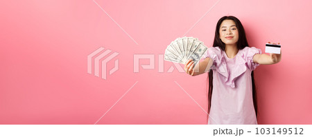 Beautiful asian woman showing plastic credit card and dollar bills, smiling pleased, standing against pink background 103149512