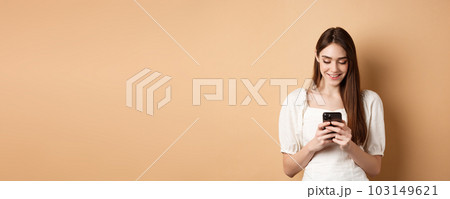 Young woman texting message on mobile phone, smiling and reading smartphone screen, standing on beige background 103149621