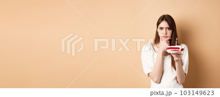 Thoughtful birthday girl frowning and thinking of wish, holding cake with candle, standing on beige background 103149623
