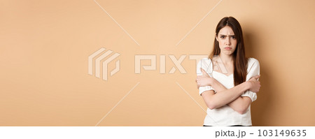 Offended woman hugging herself and frowning upset, feeling timid and defensive, standing on beige background Offended woman hugging herself and frowning upset, feeling timid and defensive, standing on beige background 103149635