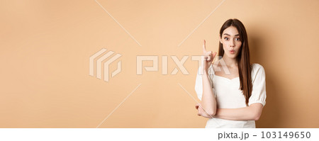 Creative girl pitching an idea, raising finger in eureka gesture, saying her plan, having a suggestion or solution, standing in dress on beige background Creative girl pitching an idea, raising finger in eureka gesture, saying her plan, having a suggestion or solution, standing in dress on beige background 103149650