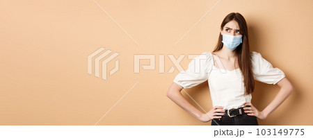 Covid-19 and lifestyle concept. Suspicious woman in medical mask frowning, looking aside at logo with doubtful face, standing on beige background Covid-19 and lifestyle concept. Suspicious woman in medical mask frowning, looking aside at logo with doubtful face, standing on beige background 103149775