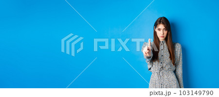 Serious woman looking confident and shaking finger, say no, stop you, disagree and disapprove, standing in dress on blue background Serious woman looking confident and shaking finger, say no, stop you, disagree and disapprove, standing in dress on blue background 103149790
