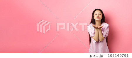 Cute and romantic asian girl blowing air kiss at camera, close eyes and smile tenderly, standing in dress against pink background Cute and romantic asian girl blowing air kiss at camera, close eyes and smile tenderly, standing in dress against pink background 103149791