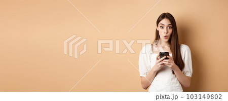 Girl standing amazed with smartphone, saying wow and look excited by awesome online offer news, standing on beige background Girl standing amazed with smartphone, saying wow and look excited by awesome online offer news, standing on beige background 103149802