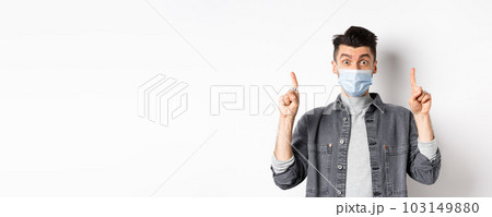 Pandemic lifestyle, healthcare and medicine concept. Excited man advertising in medical mask, pointing fingers up and look amazed, standing on white background 103149880