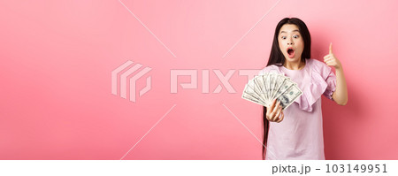 Excited teen girl holding big amount of money, showing dollar bills and thumbs up, standing amazed on pink background Excited teen girl holding big amount of money, showing dollar bills and thumbs up, standing amazed on pink background 103149951