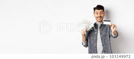 Image of handsome guy holding dollar bills but suggesting use plastic credit card, smiling friendly at camera, standing on white background 103149972