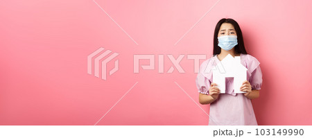 Real estate and pandemic concept. Begging asian girl showing paper house cutout, wearing medical mask, asking for apartment, standing against pink background 103149990