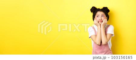 Sad moody asian girl, lean face on hands, looking left with sulking expression, standing in summer clothes on yellow background 103150165