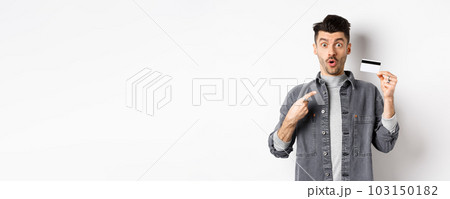 Wow look here. Excited guy pointing at plastic credit card and look amazed with awesome deal, standing against white background 103150182