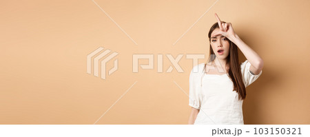 Confident woman mocking people with loser sign, being mean, standing on beige background Confident woman mocking people with loser sign, being mean, standing on beige background 103150321