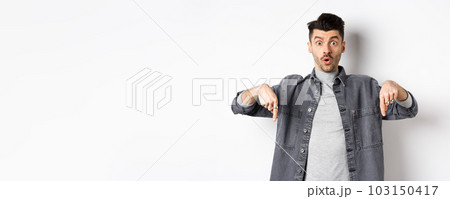 Excited guy say wow, pointing fingers down amazed, showing cool promo offer, standing on white background in denim jacket 103150417