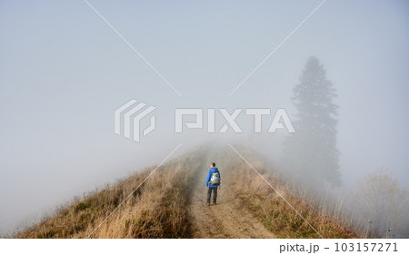 Man hiker strolling down hiking path on grassy hill with misty trees and sky in fog on background 103157271