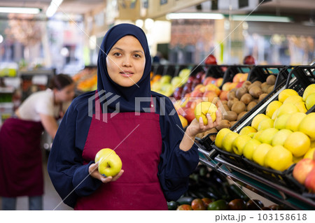 Female seller in hijab and an apron puts fresh apples on shelves in the grocery section of supermarket Female seller in hijab and an apron puts fresh apples on shelves in the grocery section of supermarket 103158618