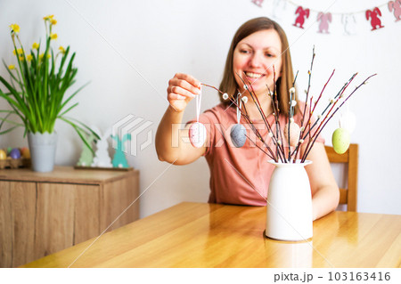 Smiling beautiful woman decorating a bouquet of willow branches with colored Easter eggs at home. Smiling beautiful woman decorating a bouquet of willow branches with colored Easter eggs at home. 103163416