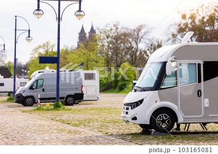 Many white modern campervan recreational motor home vehicles parked in row at camper park site Magdeburg city against Elbe river bridge. Motorhome campground stataion travel destination. RV lifestyle 103165081