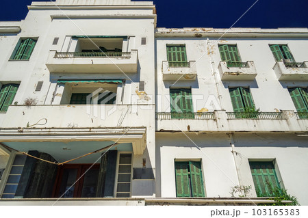 A low-rise deserted 1930s hotel with a decayed facade and green wooden window shutters under the summer blue sky. 103165383