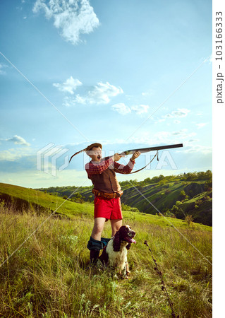 Portrait with funny hunter wearing retro hat with feather holding gun and shooting with dog English springer spaniel over nature landscape background 103166333
