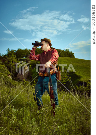 Portrait with male vintage hunter holding weapon and binoculars standing and watching, observing for prey over nature landscape background Portrait with male vintage hunter holding weapon and binoculars standing and watching, observing for prey over nature landscape background 103166383