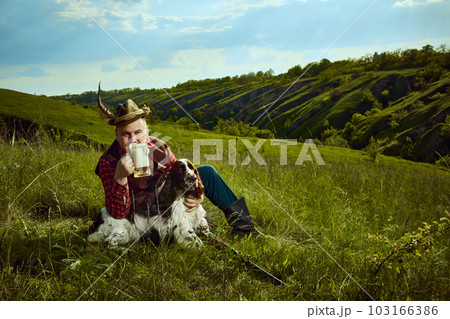 Portrait with happy man and his dog English springer spaniel sitting on grass and drinking beer after hunting on beautiful nature landscape background 103166386