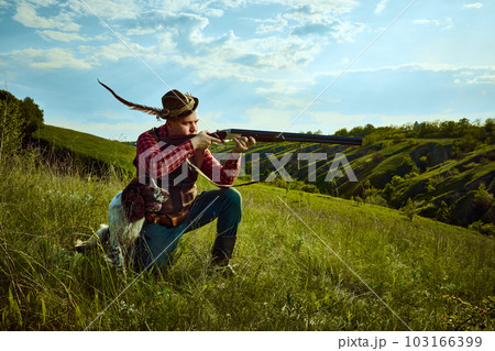 Portrait with man, vintage hunter wearing retro clothes with hat holding weapon and ready to hunt with dog English springer spaniel over nature landscape background 103166399