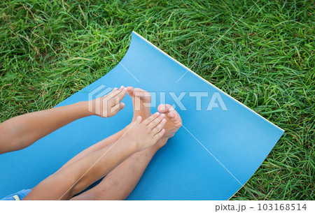 The child does exercises on a blue karemat, stretches on a rug on the grass. 103168514