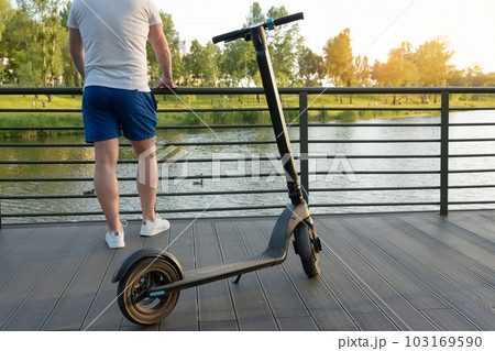 A young man stands with an electric scooter in a park near the water at sunset. Urban transport concept A young man stands with an electric scooter in a park near the water at sunset. Urban transport concept 103169590