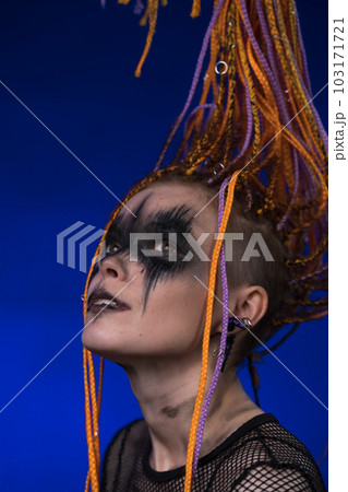 Cinematic portrait of young woman with horror black stage make-up painted on face and orange color dreadlocks hairstyle. Studio shot on blue background. Part of photo series Cinematic portrait of young woman with horror black stage make-up painted on face and orange color dreadlocks hairstyle. Studio shot on blue background. Part of photo series 103171721