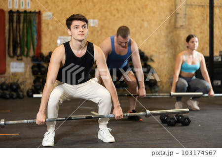 Young dedicated man doing sit-ups with rod among other people in gym 103174567