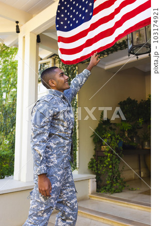 Happy biracial male american soldier wearing military uniform touching flag of usa outside the house 103174921