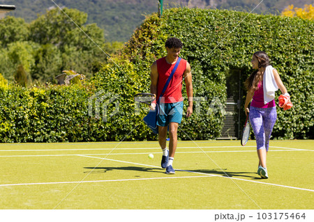 Happy diverse couple with bags and rackets walking on sunny outdoor tennis court, copy space Happy diverse couple with bags and rackets walking on sunny outdoor tennis court, copy space 103175464