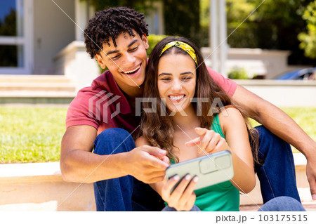 Happy diverse couple looking at smartphone and laughing sitting in sunny garden 103175515