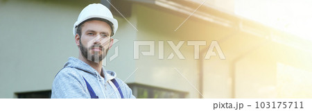 Portrait of young bearded male engineer in hard hat looking at camera, posing outdoors while working on cottage construction Portrait of young bearded male engineer in hard hat looking at camera, posing outdoors while working on cottage construction 103175711