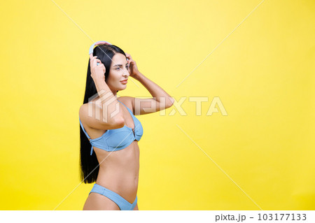 Happy young woman wearing in blue swimsuit listening music in headphones and dancing, singing song and having fun isolated on yellow wall background studio. Summer rest concept 103177133