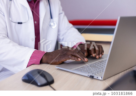 Midsection of african american male doctor sitting at desk using laptop at doctor's office Midsection of african american male doctor sitting at desk using laptop at doctor's office 103181866