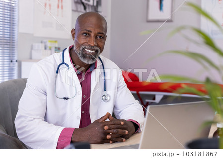 Portrait of happy african american male doctor sitting at desk and using laptop at doctor's office Portrait of happy african american male doctor sitting at desk and using laptop at doctor's office 103181867