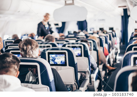 Interior of airplane with passengers on seats and stewardess in uniform walking the aisle, serving people. Commercial economy flight service concept Interior of airplane with passengers on seats and stewardess in uniform walking the aisle, serving people. Commercial economy flight service concept 103183740