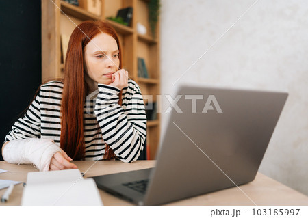 Thoughtful female freelancer with broken right hand wrapped in white gypsum bandage working on laptop computer sitting at table with paper documents. Unhealthy businesswoman working remotely from home 103185997