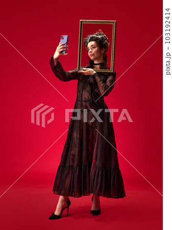 Full-length portrait of tender, beautiful, young girl holding picture frame and taking selfie with phone against red background 103192414