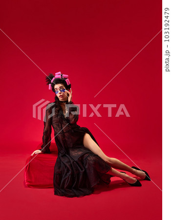 Portrait of young beautiful girl in hair curls and elegant black dress with under eye patches against red background 103192479