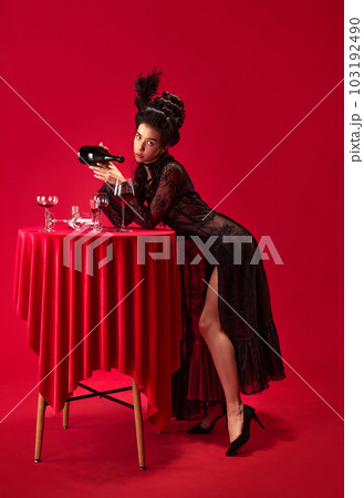 Portrait of beautiful young girl in elegant, stylish, black dress posing, pouring wine into glass against red background 103192490