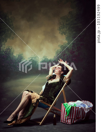 Portrait of young girl, royal person, princess in elegant dress resting, lying on chair and sunbathing against dark vintage background 103192499