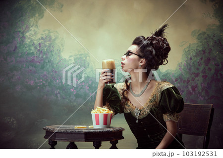 Portrait of beautiful young girl, royal person in vintage dress tasting dark foamy beer with fries against dark canvas background 103192531