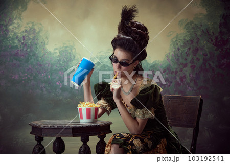 Portrait of young beautiful girl, princess, medieval royal person in elegant dress eating fries with soda against dark vintage background Portrait of young beautiful girl, princess, medieval royal person in elegant dress eating fries with soda against dark vintage background 103192541
