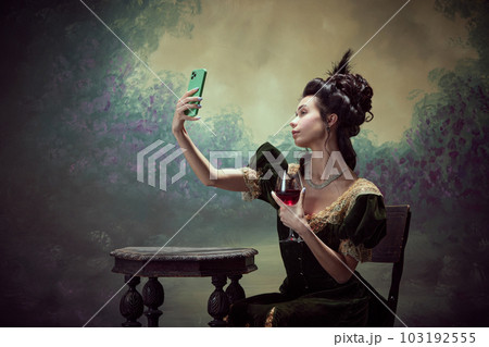Portrait of young beautiful girl, princess in elegant dress taking selfie with mobile phone and drinking red wine against dark vintage background Portrait of young beautiful girl, princess in elegant dress taking selfie with mobile phone and drinking red wine against dark vintage background 103192555