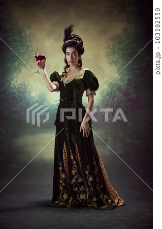Full-length portrait of beautiful young girl, princess, royal person in elegant dress standing with glass of red wine against dark vintage background 103192559