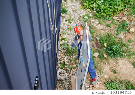 Men engineers installing solar panel system on roof of house. Workers in helmets lifting up photovoltaic solar module with help of ropes outdoors. Concept of alternative and renewable energy. 103194719